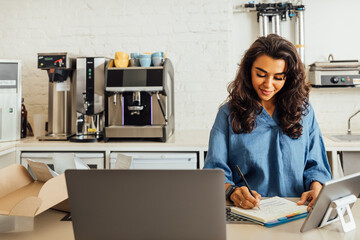 Female entrepreneur taking notes before shipping the online order to the customer. Coffee shop owner working at table.