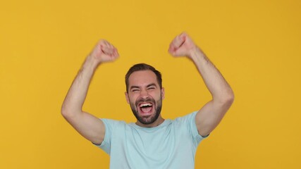 Wall Mural - Bearded fun young man 20s in basic casual blue t-shirt looking camera shocked surprised say wow yes doing winner gesture raised hands up isolated on yellow background studio. People lifestyle concept