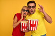 © Roman - Scared young couple two friends guy girl in 3d glasses isolated on yellow background. People sincere emotions in cinema lifestyle concept. Watching movie film, holding bucket of popcorn