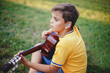 © anoushkatoronto - Hard of hearing preteen boy playing guitar outdoors. Child with hearing aids in ears playing music and singing song in park. Hobby art activity for children kids. Authentic childhood moment.