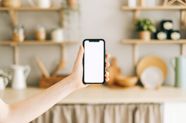 Woman hand holding smartphone with vertical white screen on kitchen at home. 