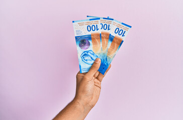 Hispanic hand holding 100 swiss franc banknotes over isolated pink background.