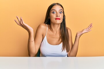 Wall Mural - Young hispanic woman wearing casual clothes sitting on the table clueless and confused with open arms, no idea and doubtful face.