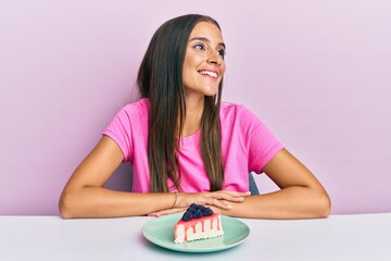 Wall Mural - Young hispanic woman eating cheesecake sitting on the table looking to side, relax profile pose with natural face and confident smile.