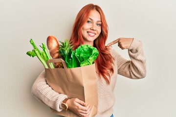 Young redhead woman holding paper bag with bread and groceries pointing finger to one self smiling happy and proud