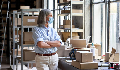 Older mature female online store small business owner, manager, stock worker, entrepreneur wearing face mask and gloves standing with arms crossed at workplace in warehouse looking through window.