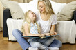 © rogerphoto - Happy family. Blonde young mother reading a book to her cute daughter while sitting at wooden floor. Motherhood concept