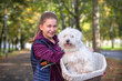 Beautiful young girl riding a bike with her cute pet white bison havanese dog in a basket through park. Autumn time.
