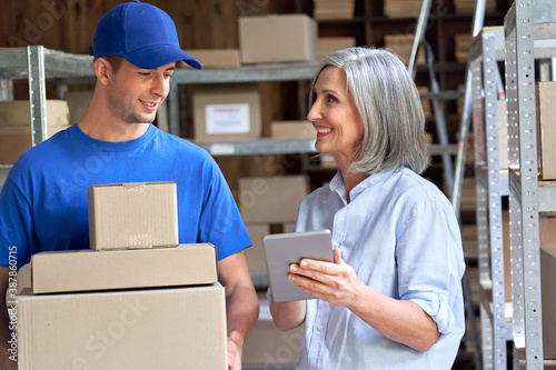 Happy mature female manager small business owner, seller, supervisor using digital tablet talking to male courier holding shipping ecommerce parcels boxes discuss delivery walking in warehouse.