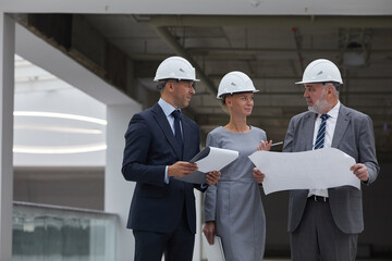 Waist up portrait of three successful business people wearing hardhats and holding plans while inspecting construction site indoors, copy space