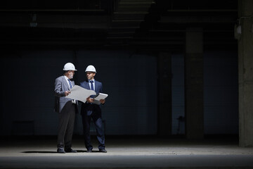 Dramatic full length portrait of two mature business people wearing hardhats and holding plans while standing in dark at construction site lit by harsh lighting, copy space