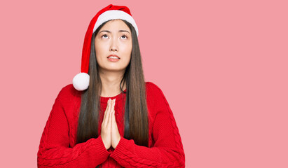 Young chinese woman wearing christmas hat begging and praying with hands together with hope expression on face very emotional and worried. begging.