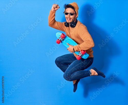 Young handsome latin man wearing wool cap and thug life glasses.  Jumping with smile on face holding skate over isolated blue background