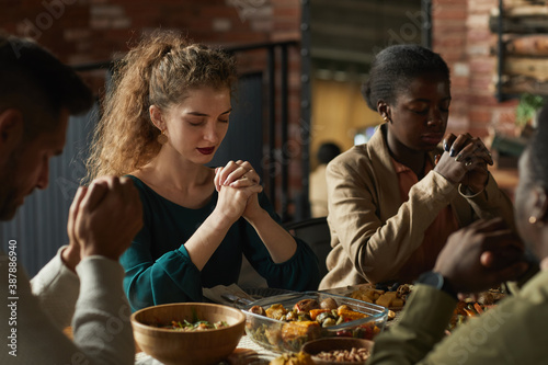 Warm toned portrait of multi-ethnic group of elegant young people praying with eyes closed while sitting at dinner table during Thanksgiving celebration, copy space
