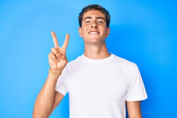 Wall Mural - Young handsome man wearing casual white t shirt smiling with happy face winking at the camera doing victory sign. number two.