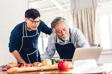 Wall Mural - Portrait of happy love asian family senior mature father and young adult son having fun cooking together and looking for recipe on Internet with laptop computer to prepare the yummy eating lunch 