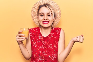 Young beautiful blonde woman wearing summer hat drinking glass of orange juice screaming proud, celebrating victory and success very excited with raised arm