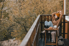 Young Woman In Knitted Sweater And Hat Drinking Tea And Eating Fresh Croissants On Cozy Balcony Of A Wooden Country House.