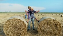 Parenting Pleasures, Happy Father Spends Time Playing With Baby On Straw Bales In Field, Male Child Jumping On Bales With Help Of Father During Family Outing Outdoors