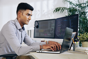 Sticker - Pensive young Indian programmer sitting at office desk with two monitors and typing on laptop