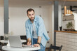 © ViDi Studio - Confident handsome young man standing alone near table in coffee shop cafe restaurant indoors working or studying on laptop pc computer writing in notebook. Freelance mobile office business concept.