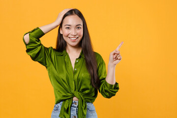 Wall Mural - Smiling cheerful young brunette asian woman wearing basic casual green shirt standing put hand on head pointing index finger up on mock up copy space isolated on yellow background, studio portrait.