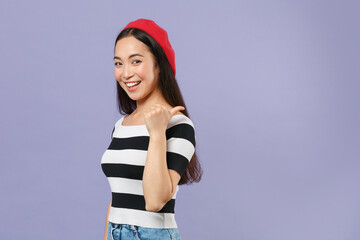 Poster - Side view of smiling cheerful young asian woman 20s wearing striped t-shirt red beret standing pointing thumb aside on mock up copy space isolated on pastel violet colour background studio portrait.