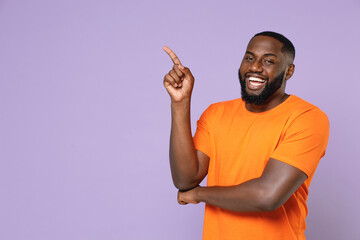 Wall Mural - Smiling cheerful young african american man 20s wearing basic casual orange t-shirt standing pointing index finger up on mock up copy space isolated on pastel violet colour background studio portrait.