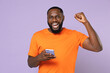 © ViDi Studio - Happy young african american man 20s wearing basic casual orange t-shirt doing winner gesture using mobile cell phone typing sms message isolated on pastel violet colour background, studio portrait.