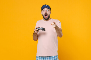 Poster - Shocked young man in pajamas home wear sleep mask play pc game with joystick console pointing index finger on camera resting at home isolated on yellow background. Relax good mood lifestyle concept.