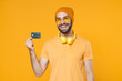 © ViDi Studio - Smiling cheerful young bearded man 20s wearing basic casual t-shirt headphones eyeglasses hat standing hold in hand credit bank card looking camera isolated on yellow background, studio portrait.
