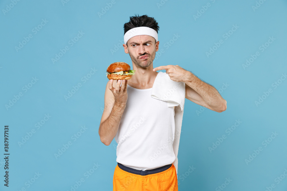 Confused young sporty man with skinny body sportsman in headband shirt towel point index finger on american classic fast food burger isolated on blue background. Workout gym sport motivation concept. - obrazy, fototapety, plakaty 