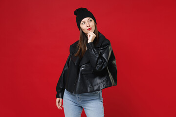 Beautiful attractive pensive young brunette woman wearing casual black leather jacket white t-shirt hat put hand prop up on chin looking aside isolated on bright red colour background studio portrait.