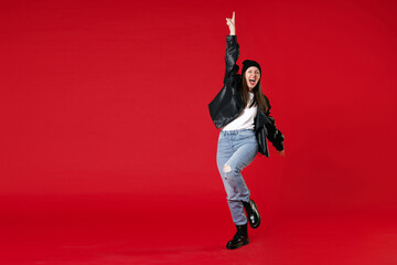 Wall Mural - Full length of screaming laughing cheerful young brunette woman 20s in black leather jacket white t-shirt hat pointing index finger up dancing isolated on bright red colour background studio portrait.