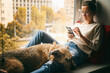© olezzo - A young woman with her brown dog is sitting on a windowsill overlooking the city, looking at the smartphone screen and stroking her pet
