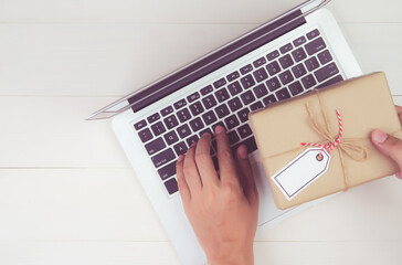 Wall Mural - Hand of man holding gift box and tag having laptop computer on wooden table, present giving for Christmas day or birthday, season and celebrate, top view, flat lay, online shopping concept.