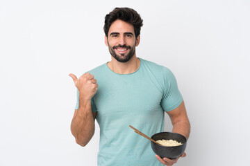 Wall Mural - Young handsome man with beard over isolated white background pointing to the side to present a product while holding a bowl of noodles with chopsticks