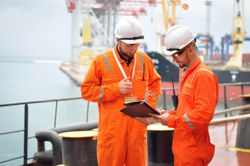 Two sailors in orange overalls work on the ship with documents.