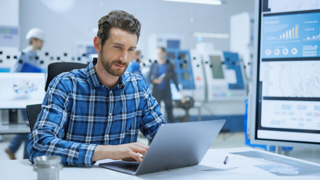 Modern Industrial Factory: Portrait of Industry Engineer Sitting at His Desk, Working on Laptop, Analyzing Mechanism and Blueprints. Background Functional Manufactory with Working on CNC Machinery