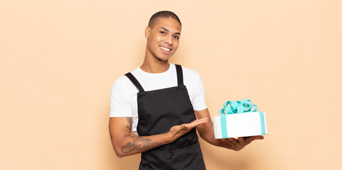 Wall Mural - young black man smiling cheerfully, feeling happy and showing a concept in copy space with palm of hand