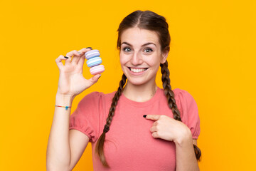 Wall Mural - Young woman over isolated yellow background holding colorful French macarons with surprise expression