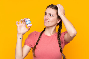 Wall Mural - Young woman over isolated yellow background holding colorful French macarons with confuse expression