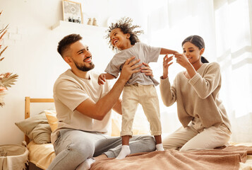 Wall Mural - happy diverse family mom, dad and child  laughing, playing and jumping   in bed   at home.