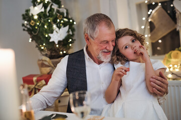 Wall Mural - Senior grandfather with small granddaughter indoors at Christmas, sitting at table.