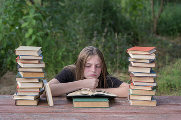 Young girl reading a book and dreams in the garden at a wooden table with a stack of books.