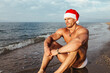 © Miguel Frias/Westend61 - Thoughtful shirtless young man wearing Santa hat sitting at beach