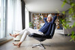 © Rainer Berg/Westend61 - Contemplating senior woman with hands behind head sitting on chair at home
