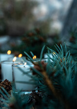 Five Lit White Votive Candles Burning Flames Amid Green Pine Branches And Decorative Pine Cones