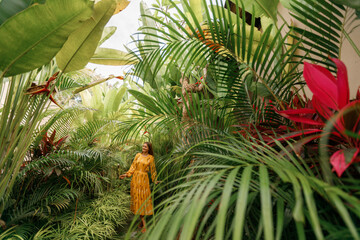 Ypoung happy woman walking in tropical garden in long yellow dress. Brunette with long dark hair  enjoying nature during vacation on Bali tropical island