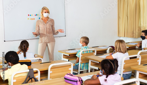 Young female teacher in protective face mask giving lesson to children in elementary school. New life reality during coronavirus pandemic
